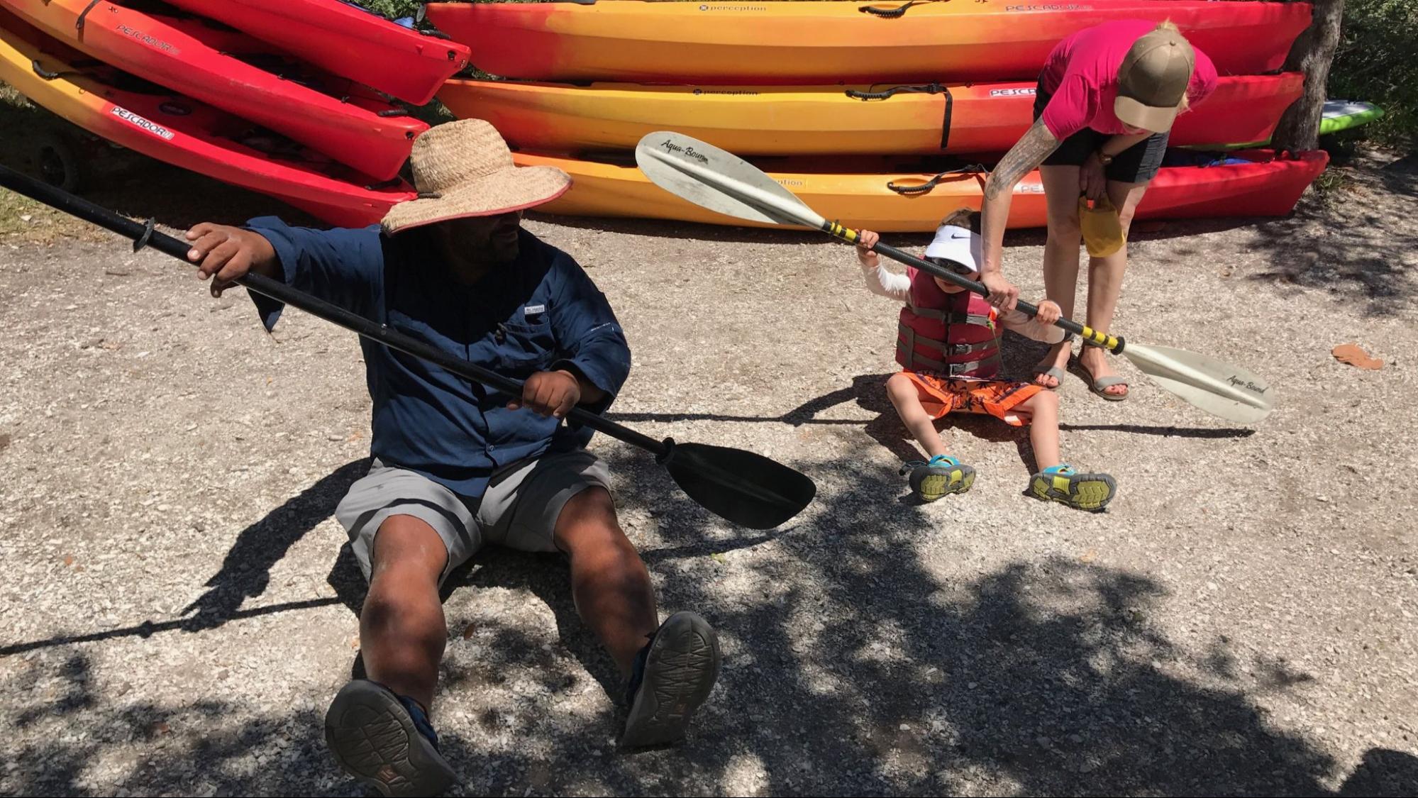 An expert guide from Paradise Adventures sitting on the shore, patiently showing a young child in a life vest how to hold a kayak paddle before hitting the water. Bright orange tandem kayaks are stacked in the background.