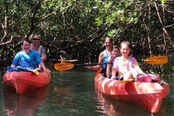 A happy family of five smiling while paddling through a shaded, lush green mangrove tunnel in bright orange tandem kayaks.