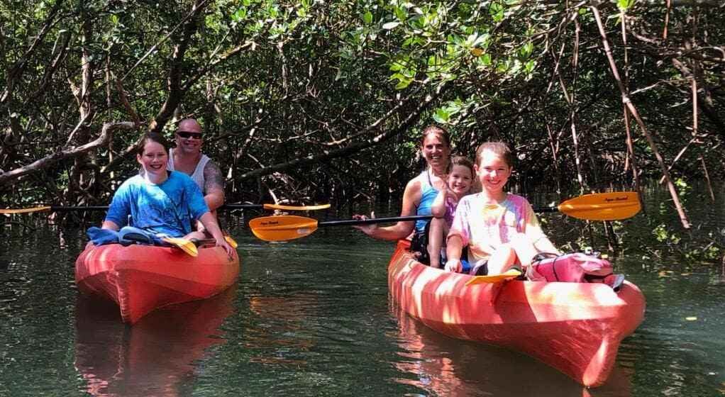 A happy family of five smiling while paddling through a shaded, lush green mangrove tunnel in bright orange tandem kayaks.