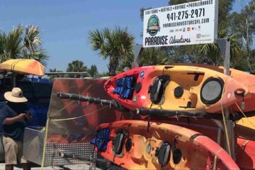 The Paradise Adventures Sarasota launch area, showing colorful kayaks stacked on trailers, a truck, and a large sign with the company logo and contact phone number.