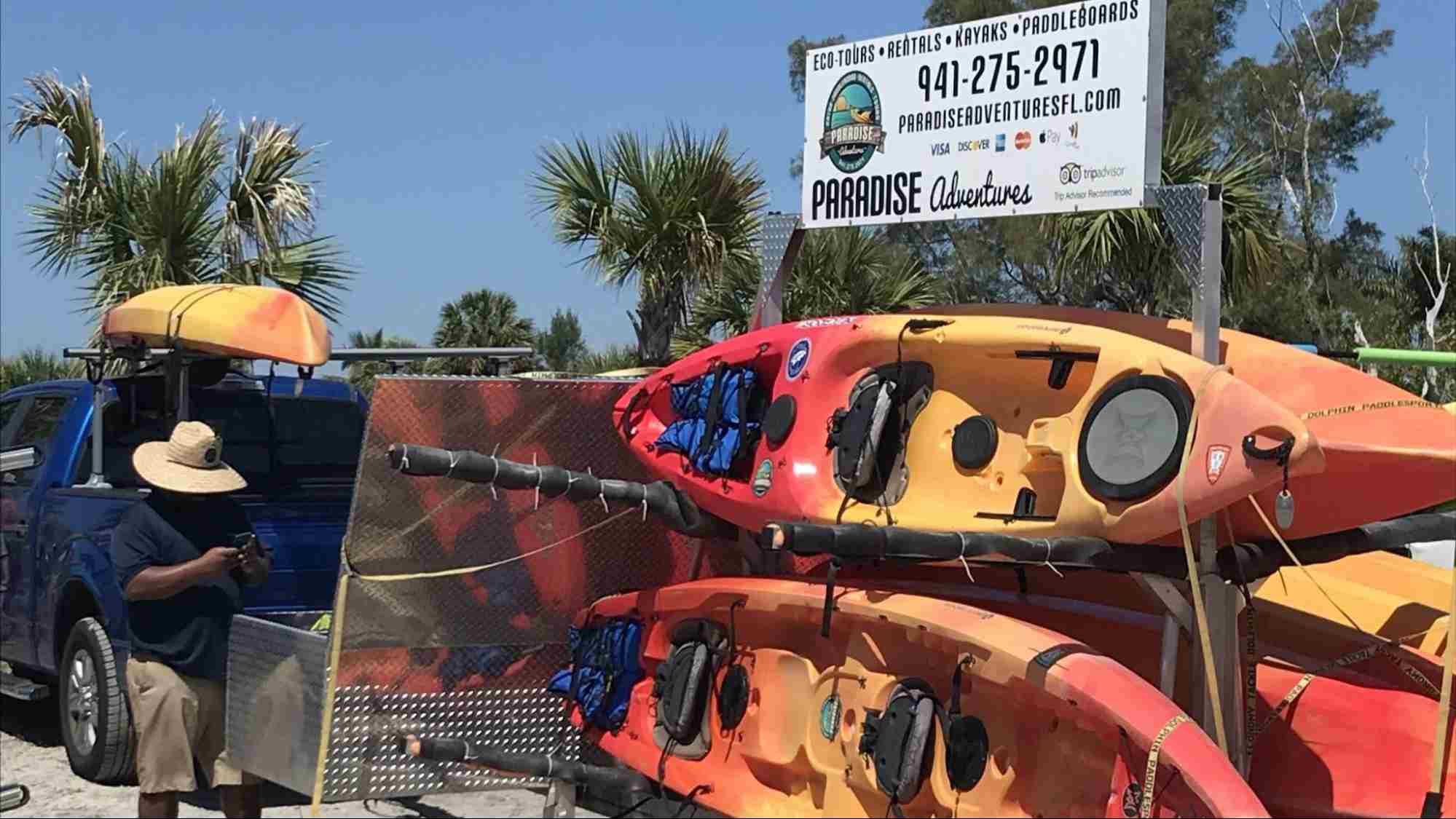 The Paradise Adventures Sarasota launch area, showing colorful kayaks stacked on trailers, a truck, and a large sign with the company logo and contact phone number.